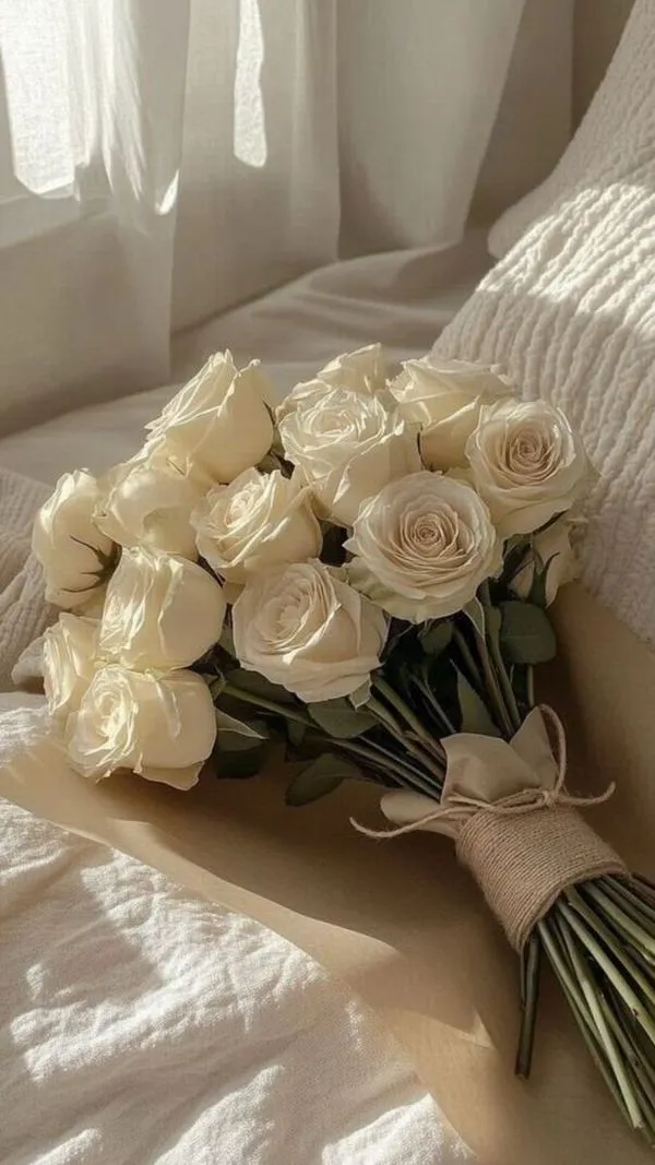 Bó hoa hồng trắng đặt trên giường, ánh sáng mềm qua rèm, cảm giác dịu dàng và ấm áp