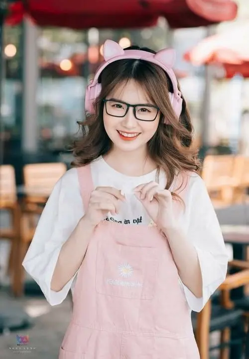 48 Cô bạn đeo kính mặc áo hồng nhạt đứng giữa không gian quán ngoài trời, hai tay giữ ly trước ngực và cười tươi, ảnh k5 nhìn rất cute và thân thiện