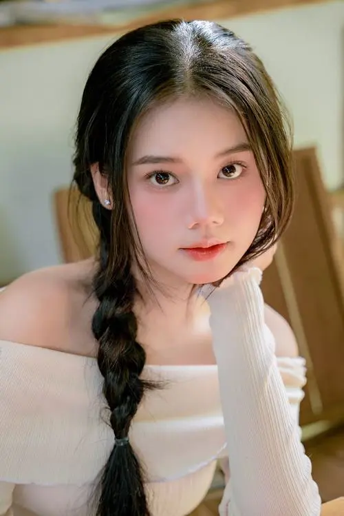 32 Cô nàng buộc tóc hai bên mặc áo trễ vai sáng màu, chống tay lên bàn và nhìn thẳng máy, chân dung 2k5 này nhỏ nhắn và cute như ảnh chụp phòng ngủ