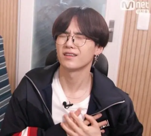 24 Chàng trai tóc đen ngồi trong khung phỏng vấn, tay giữ ngực và cười ngượng, áo tối màu cùng nền ấm làm meme đau tim này trông khá gần gũi