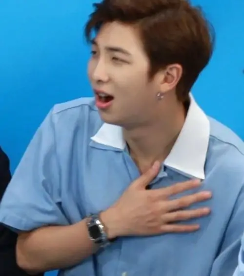 27 Nam thần mặc vest xanh ngồi trên nền sân khấu sáng, một tay đặt trước ngực và mặt hơi nhăn lại như đang diễn cú nhồi máu cơ tim ảo khá ngọt