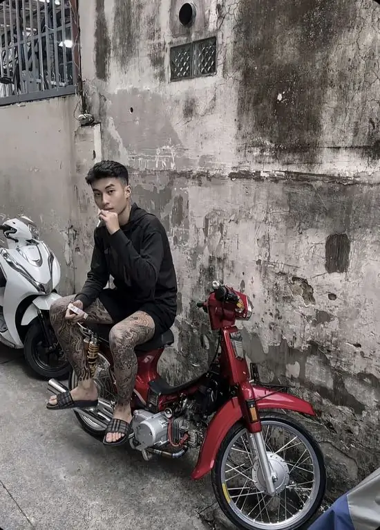 7 Hình profile nam thanh niên ngồi trên chiếc xe Cub cũ màu đỏ, trang phục giản dị nhưng toát lên vẻ phong trần của một kẻ bụi đời.
