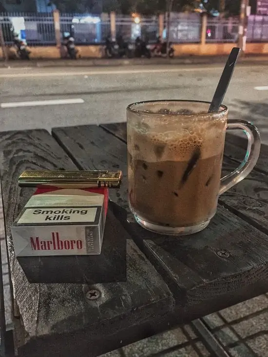 9 Khoảnh khắc bàn ngoài trời ban đêm với ly cà phê đá và gói thuốc lá, tay cầm điếu thuốc đang hút, mang vẻ suy tư.