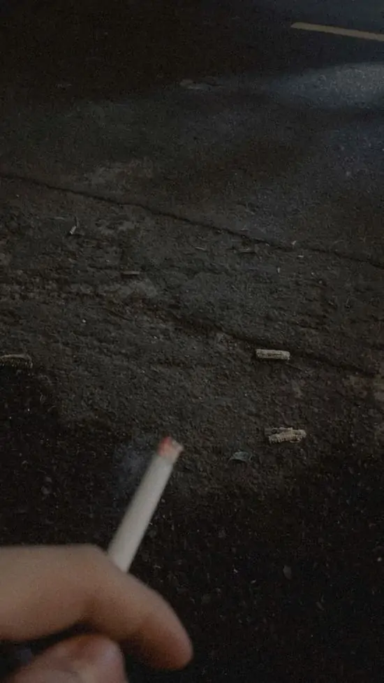 12 Bàn tay cầm điếu thuốc gần sát, tro rơi nhẹ, nền đen hoàn toàn trong hình ảnh hút thuốc u buồn.