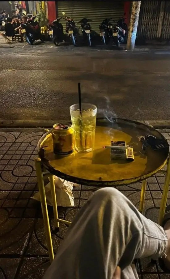 1 Ảnh đại diện chàng trai ngồi ngoài trời đêm bên ly nước vàng trên bàn nhỏ, chân bắt chéo thư giãn, khói thuốc bay nhẹ mang vẻ u uất.
