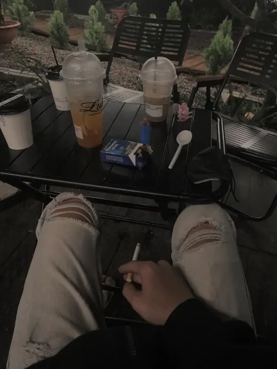 26 Chàng trai ngồi trên ghế dài trong phòng, chân duỗi ra bàn, cầm điếu thuốc hút, khói bay chìm ngập.