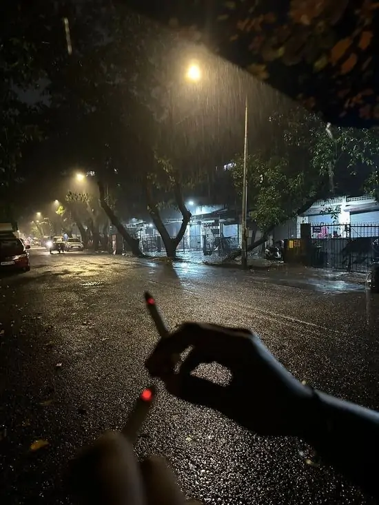 3 Hình profile tay cầm điếu thuốc trên mặt đường ướt át ban đêm, ánh đèn đường phản chiếu xuống vũng nước, trông rất trầm ngâm.