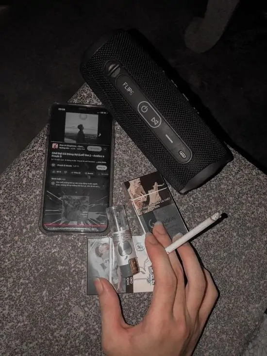 8 Tay cầm điếu thuốc trên bàn với điện thoại và chai đen, khói thuốc lan tỏa chậm rãi, cảm giác lạnh lùng.