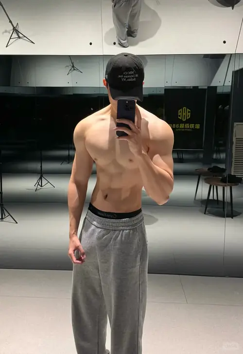 27 Trước gương phòng gym, chàng trai đội mũ đen cầm điện thoại, quần xám và thân hình gọn chắc