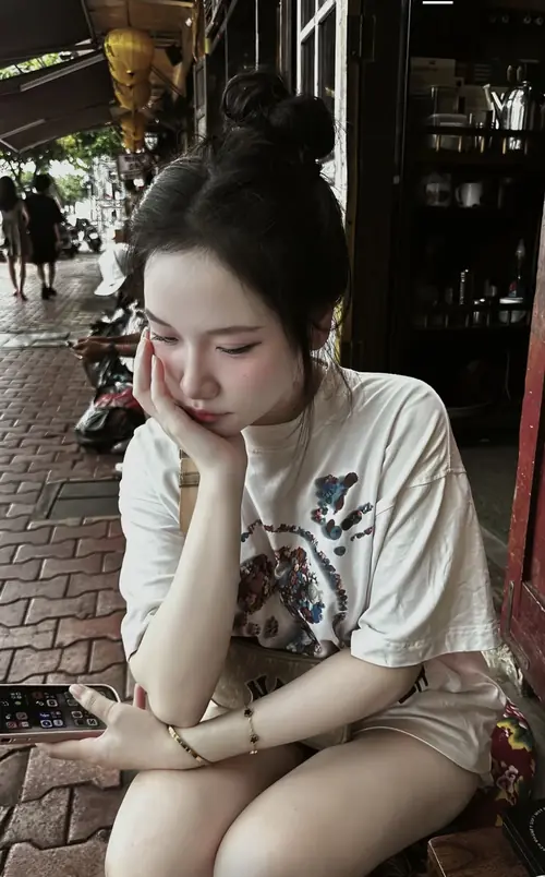 7 Cô nàng tóc búi ngồi trước quán, áo thun trắng rộng và gương mặt nghiêng nhẹ khá trầm