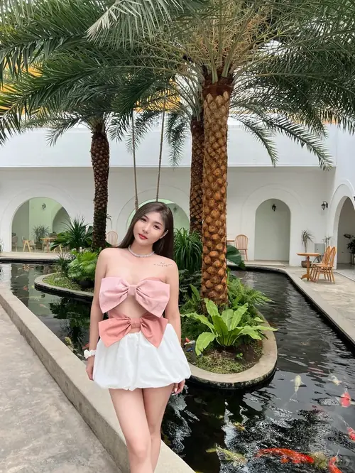 2 Giữa sân nghỉ dưỡng có hàng cọ, cô gái mặc áo hồng váy trắng đứng cạnh hồ nước