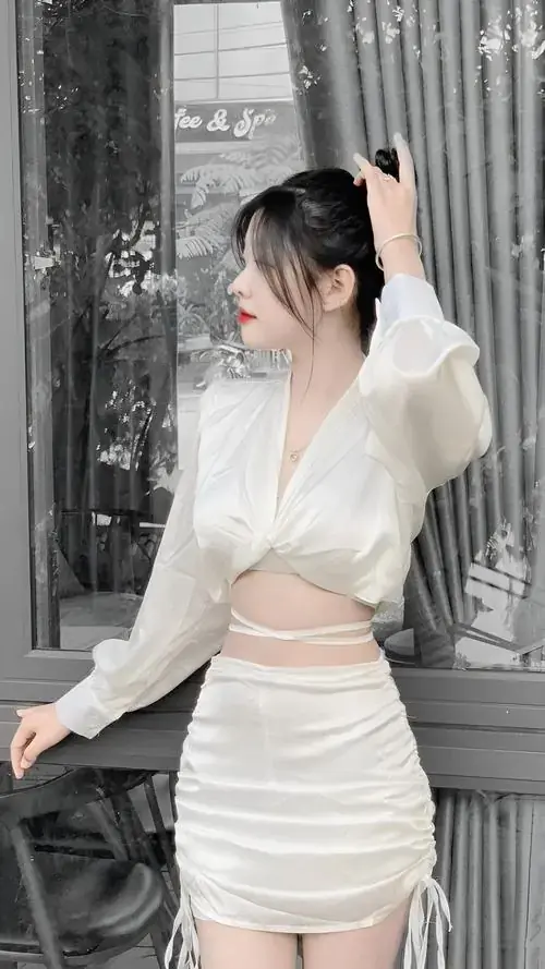 13 Nàng buộc tóc cao mặc áo trắng tay dài và chân váy ngắn, ngồi cạnh khung cửa xám