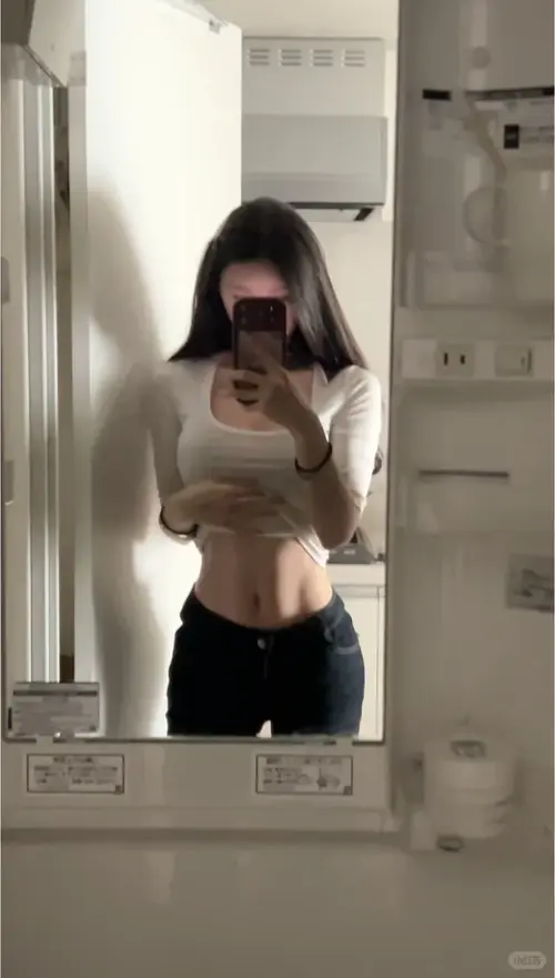 12 Trong phòng tập nhiều gương, cô gái mỉm cười nhẹ, áo đen ngắn và quần xám ôm.