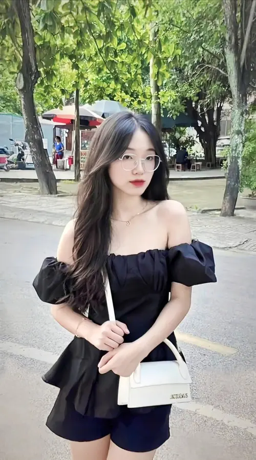 1 Cô nàng mặc áo trễ vai đen đứng dưới hàng cây, tóc xoăn dài và mắt kính mảnh rất duyên