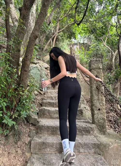 1 Cô gái đứng trên bậc đá rợp cây, tóc xõa, áo sát nách và quần ôm đen.
