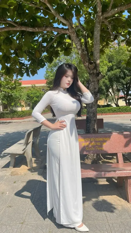 2 Dưới bóng cây có ghế đá, cô gái mặc áo dài trắng đưa tay chỉnh tóc và nhìn xuống nhẹ.