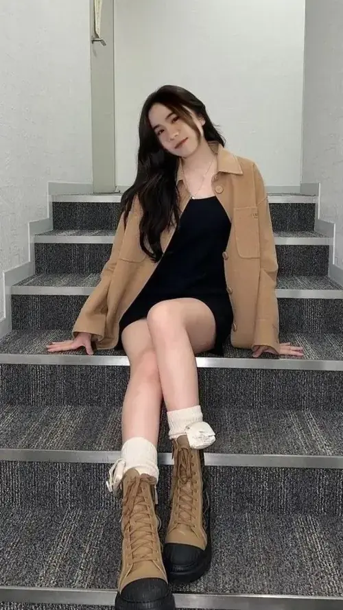 2 Trên bậc thang xám, nữ streamer Hero Team ngồi nghiêng với áo đen, blazer nâu và boots cao cổ