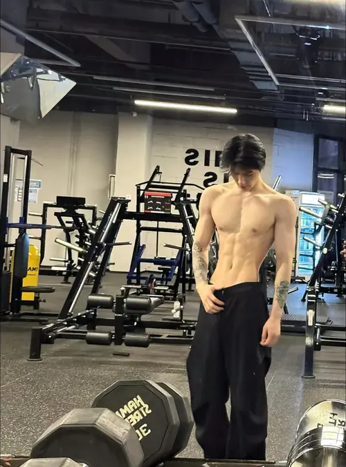 2 Trong phòng gym nhiều máy tập, chàng trai quay lưng khoe lưng trần, tóc tối màu buông tự nhiên