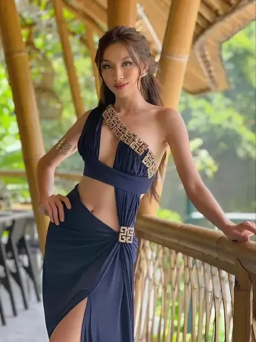 17 Nguyễn Thúc Thùy Tiên đứng bên lan can gỗ, trang phục đen phối họa tiết vàng và nụ cười duyên