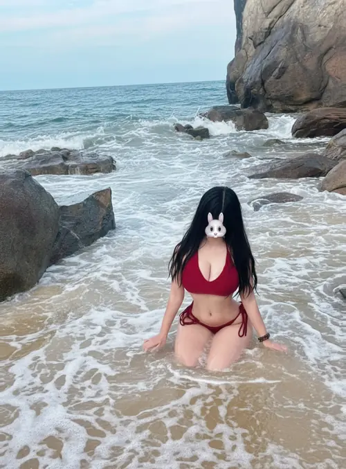 15 Cô nàng diện bikini đỏ ngồi giữa bọt sóng, phía sau là biển và đá lớn.