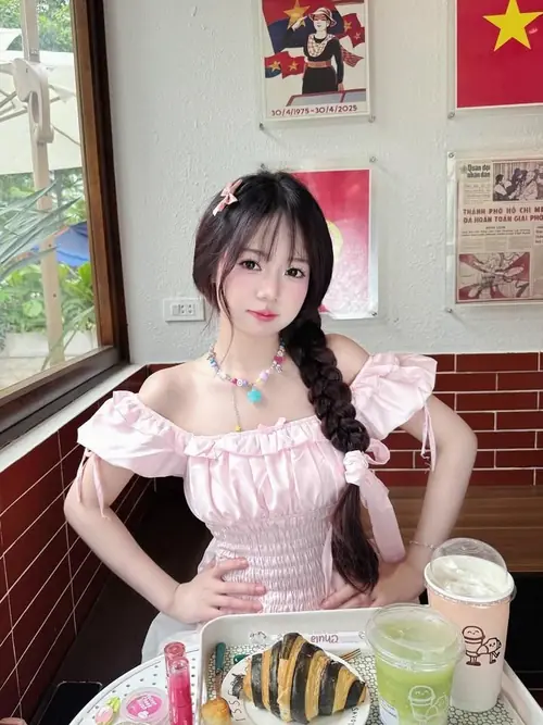 23 Cô gái váy hồng tay phồng đứng trong quán, tóc ngắn ôm mặt và hai ly đặt trước bàn