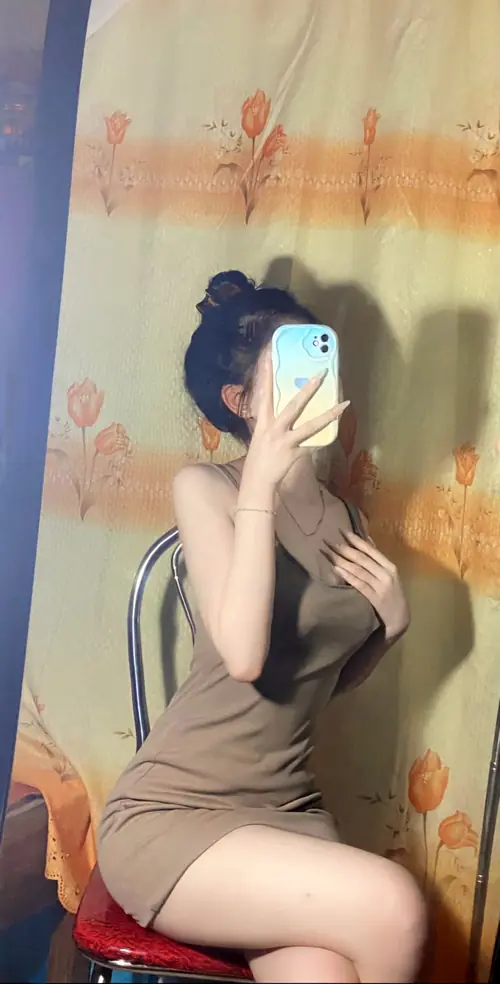17 Trước gương phòng tắm, cô gái mặc váy nâu ôm sát ngực, ngồi co chân cầm điện thoại
