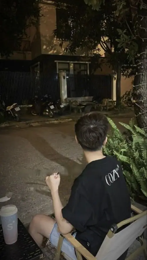17 Cậu bạn ngồi quay lưng trong khoảng sân đêm, áo đen giản dị và khung cảnh nhiều cây