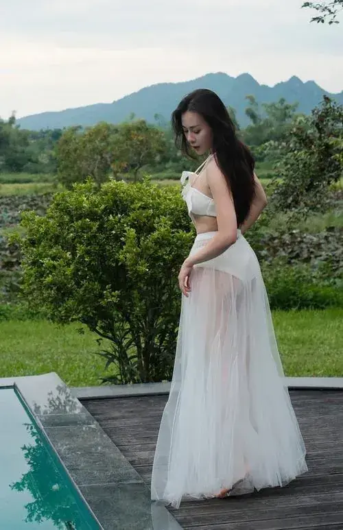 11 Người đẹp mặc váy trắng đứng cạnh hàng cây xanh, tóc dài buông sau lưng và nhìn nghiêng rất dịu